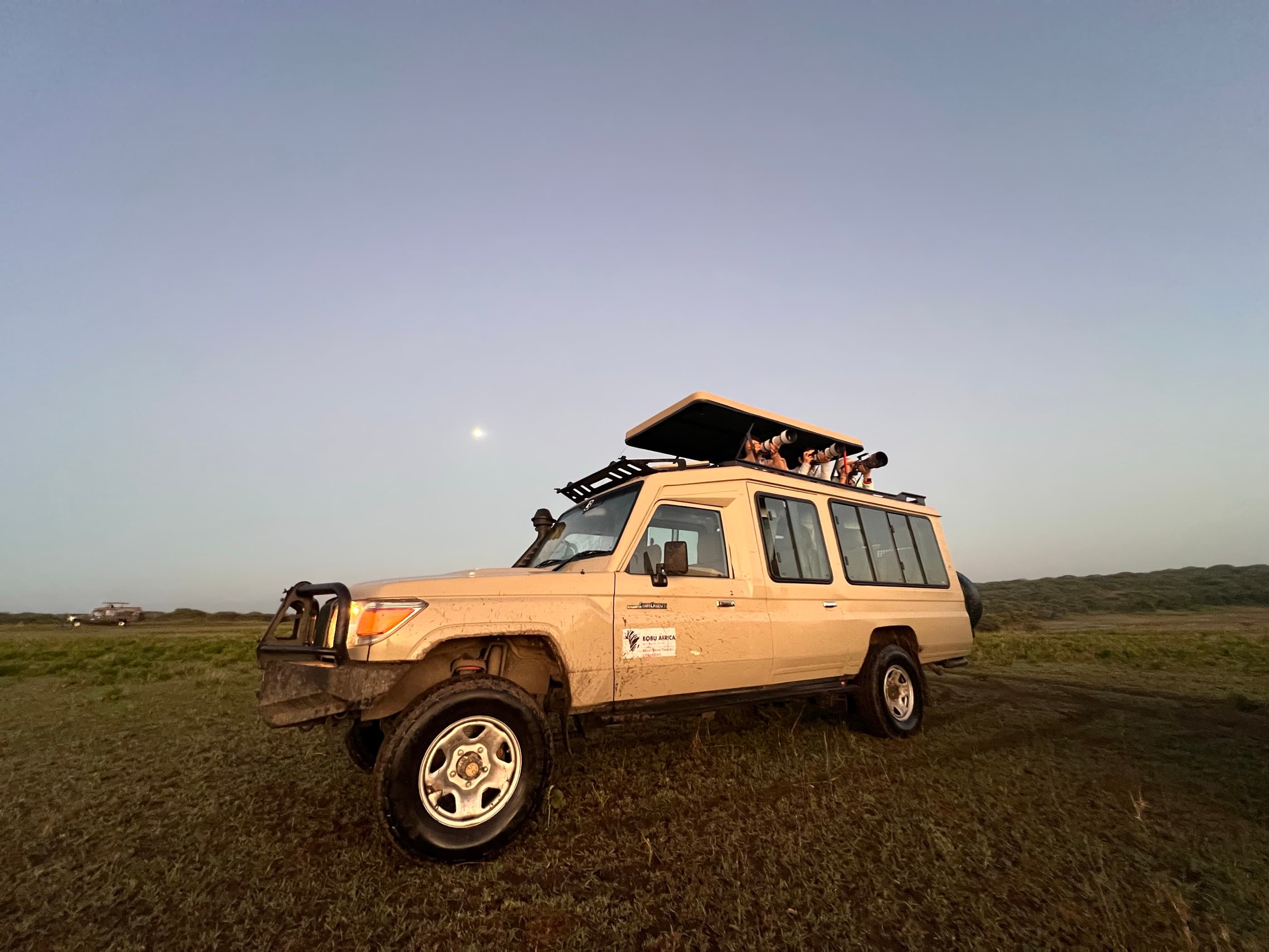A fully enclosed Toyota Land Cruiser with its pop-up roof open, photographers visible inside shooting with long telephoto lenses, on open grassland at dusk — a second vehicle parked in the far distance.