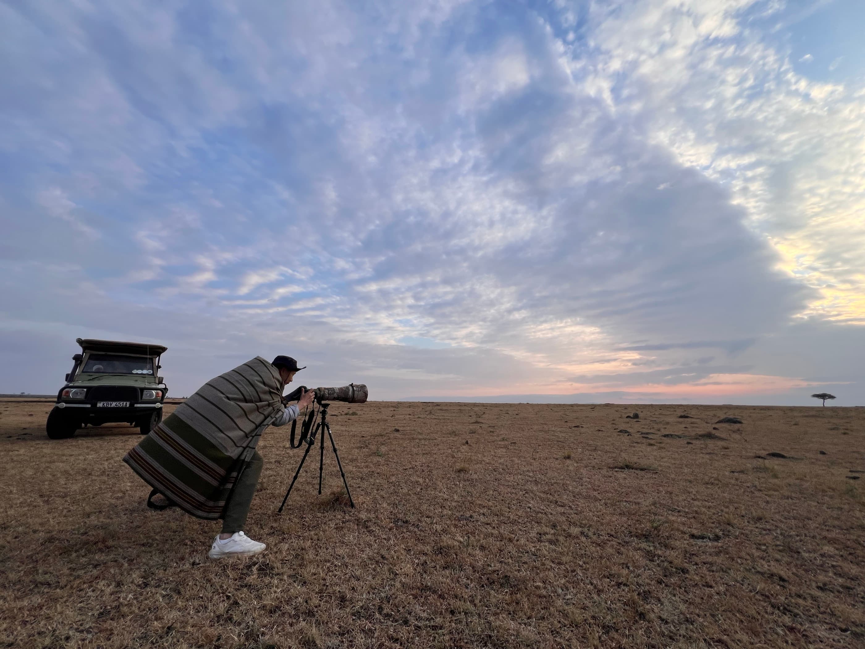 A photographer wrapped in a striped blanket, crouched at a tripod-mounted long lens on the open Mara plains at dawn, the vehicle parked behind them, a vast pale sky overhead.