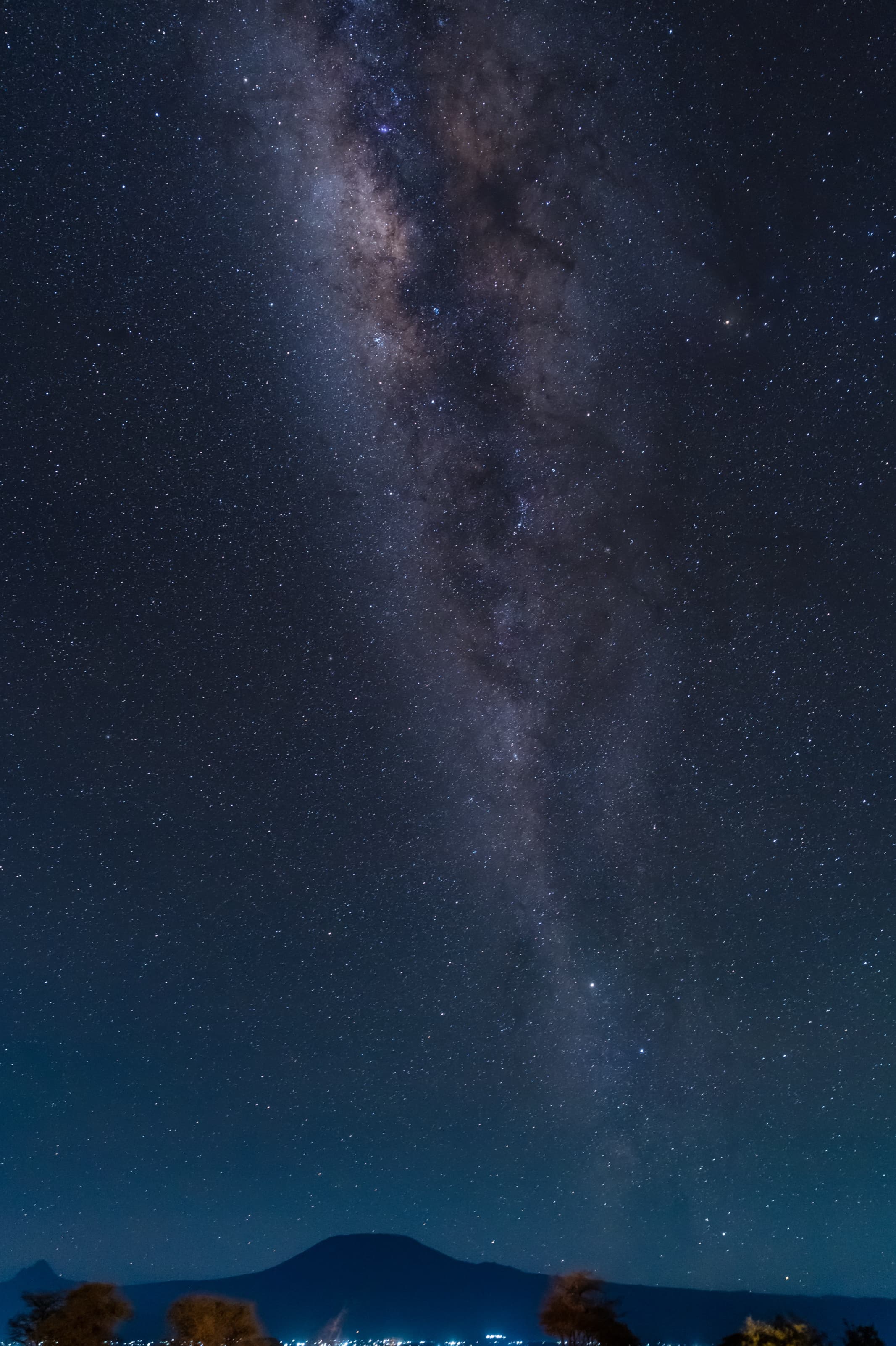 The Milky Way rising as a bright band of stars above the dark silhouette of Kilimanjaro at night, with faint lights along the base of the mountain.