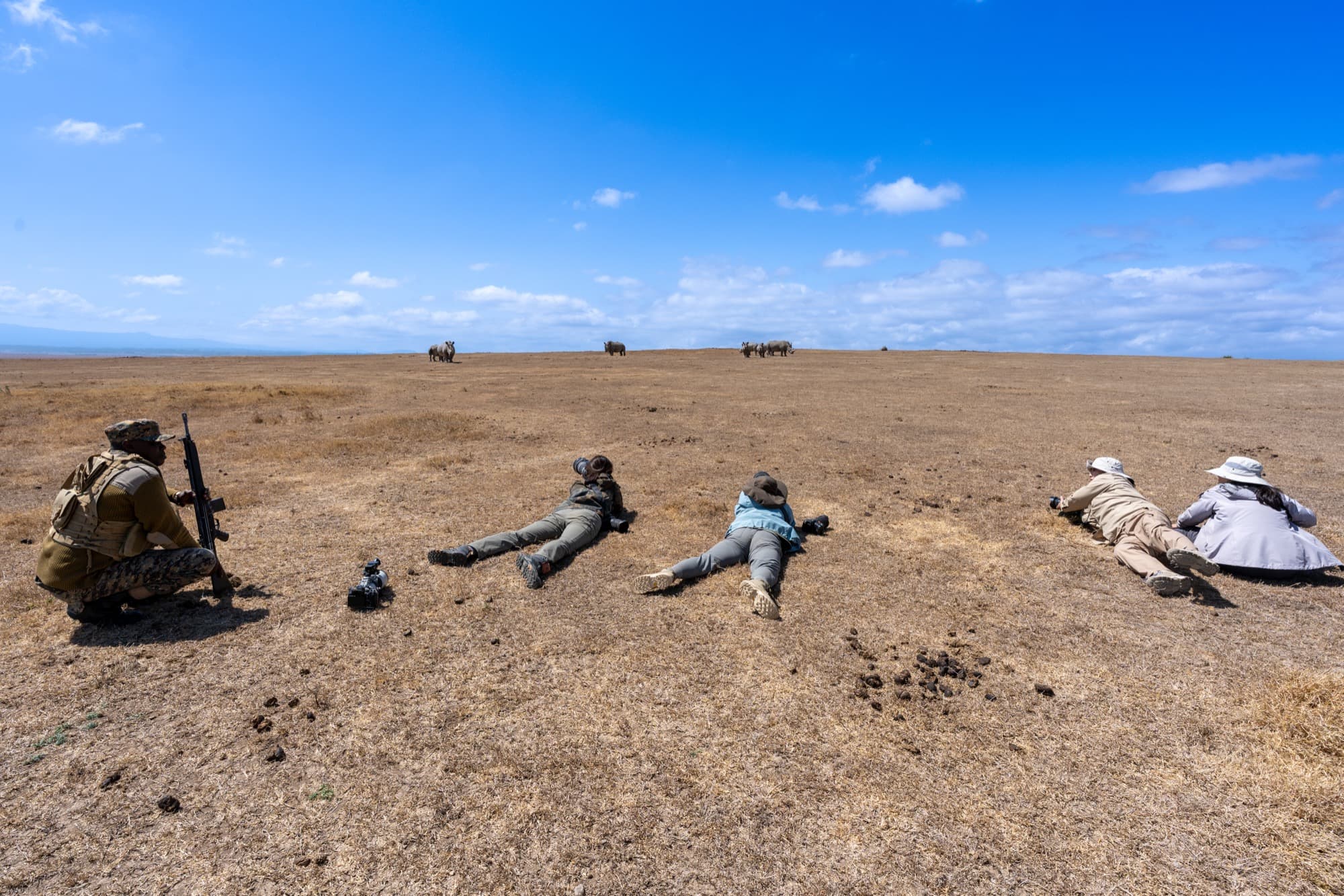 Four photographers prone in a line, cameras at earth level — three white rhinoceros on the ridge behind them, a KWS ranger standing to one side.